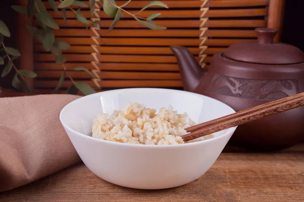 Riz brun cuit dans un bol blanc avec des baguettes sur le fond en bois . — Photo