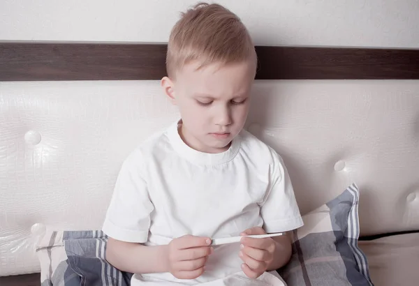 Niño enfermo sentado en la cama y mirando el termómetro digital. El chico malo está midiendo la temperatura corporal y no se siente bien —  Fotos de Stock