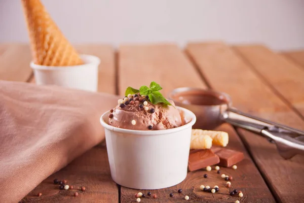 Paper bowl of chocolate ice cream, waffle cone and spoon for ice cream on wooden background.