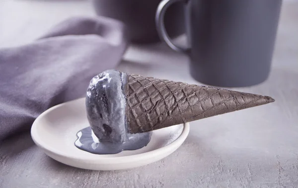 Black ice cream in traditional portioned ice cream cones. On a white ceramic plate, on a gray table with black mug and gray napkin.