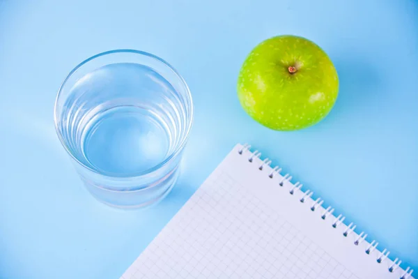 Manzana fresca, vaso de agua y cuaderno sobre fondo azul . — Foto de Stock