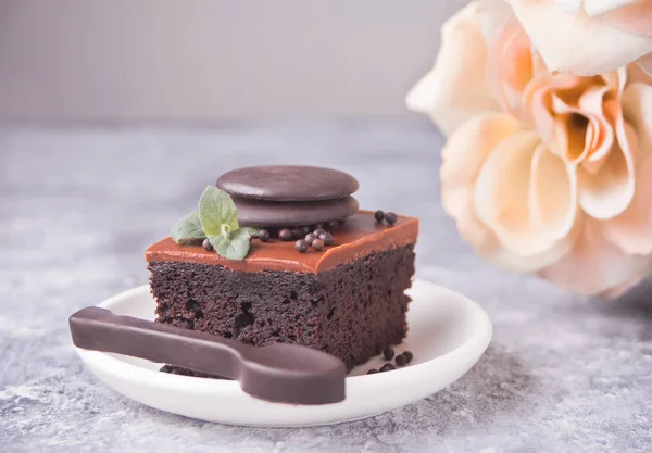 A piece of homemade chocolate cake on the plate with icing, mint leaf and chocolate spoon on the gray table — Stock Photo, Image