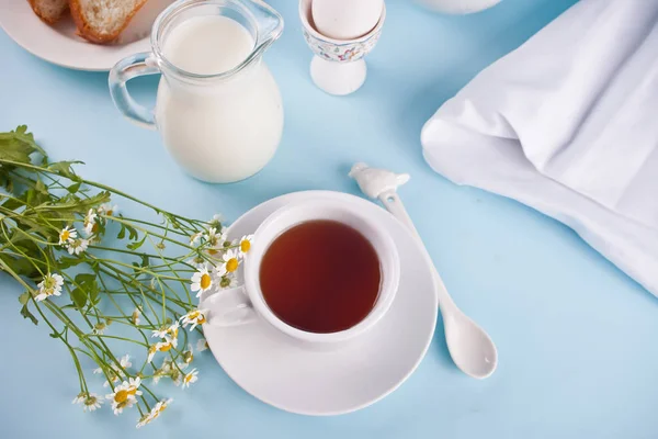 Bom dia. Pequeno-almoço. Xícara de chá com leite, jarra com leite, ovo no fundo azul — Fotografia de Stock
