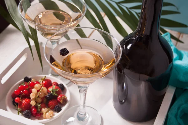 Witte wijn in een glazen, fles en bord met bessen op de witte houten dienblad onder het palmtak. Diner voor twee. — Stockfoto