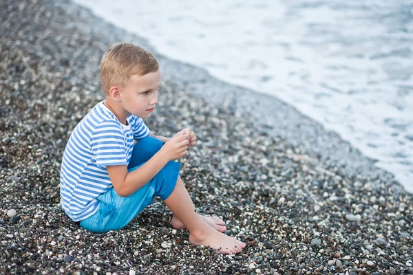 Pojke i en randig t-shirt som sitter i närheten av vattnet på stranden — Stockfoto