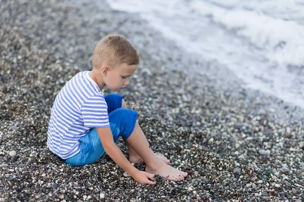 ビーチの水辺に座っているストライプのTシャツを着た少年 — ストック写真