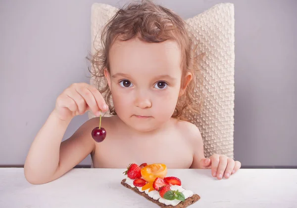 Маленькая милая девочка держит вишню в руке и ест хрустящий хлеб со сливочным сыром, фруктами и ягодами . — стоковое фото