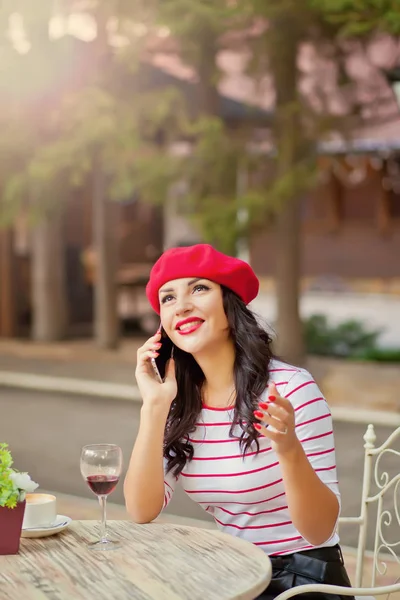 Mulher em um boné vermelho bebendo vinho tinto no café ao ar livre e falando ao telefone — Fotografia de Stock