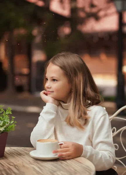 Hezká holčička se usmívá a pije cappuccino ve venkovní kavárně — Stock fotografie