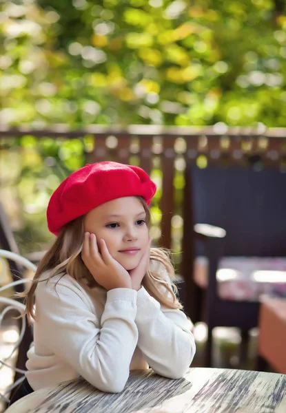 Ładna dziewczynka w czerwonym kapelusza siedzi w kawiarni na świeżym powietrzu i marzy — Zdjęcie stockowe