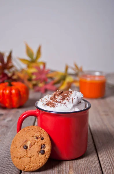 Красная кружка горячего сливочного какао с пеной на деревянном столе с печеньем, осенними листьями, свечами и тыквой на заднем плане — стоковое фото