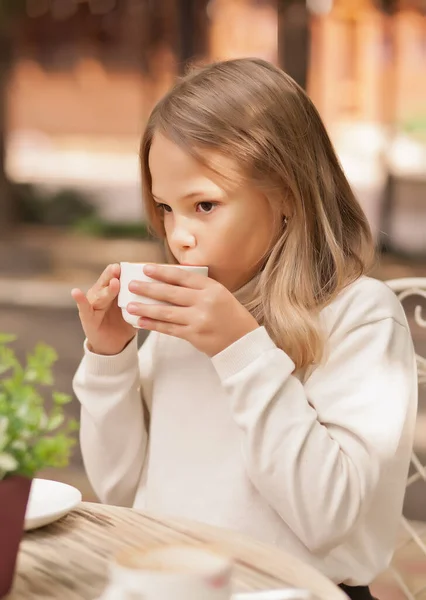Красивая маленькая девочка улыбается и пьет капучино в кафе на открытом воздухе — стоковое фото