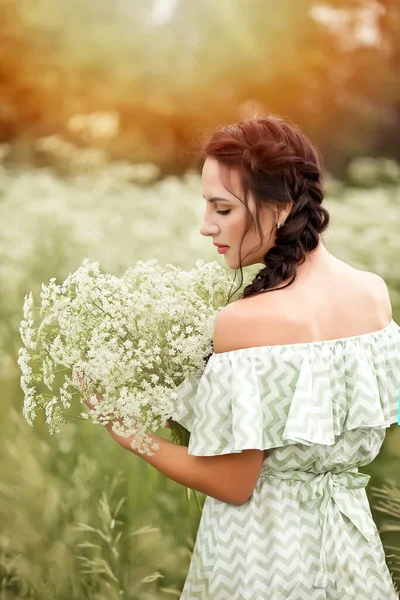 Beautiful young brunette woman in a green dress on the meadow with white flowers in a hand on a warm summer day — Stock Photo, Image