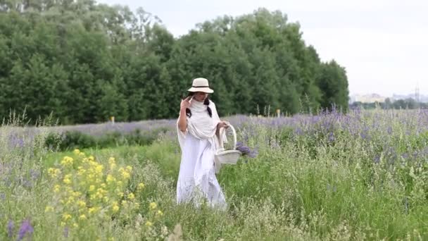 Žena v klobouku kráčí po louce a sbírá šeříkové květy v proutěném koši.