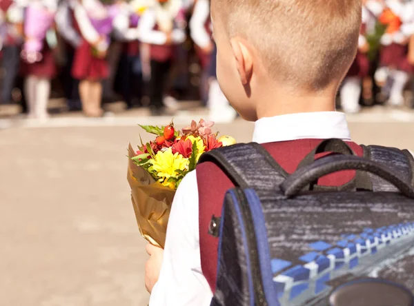 リュックと花を持つ小さな男の子は学校に行きます。バックビュー. — ストック写真