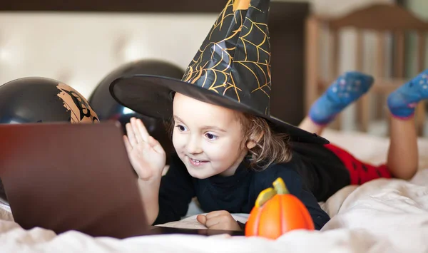 ハロウィーンの魔女の衣装を着た面白い子供の女の子はベッドの上にあり デジタルタブレットノートパソコンのノートブックを使用しています オンライン通話の友人や両親 ロイヤリティフリーのストック画像