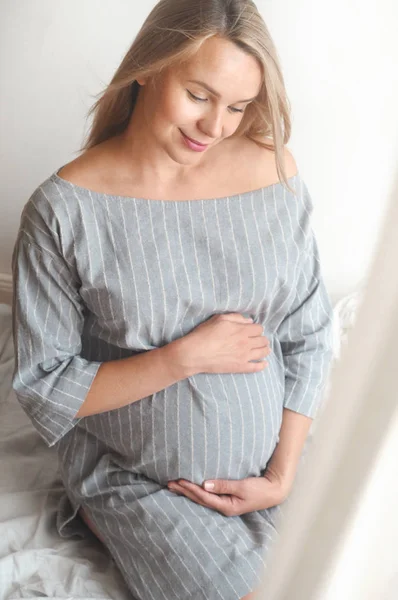 Boldog, terhes nő ül egy ágyban otthon, és keresi a kamera — Stock Fotó
