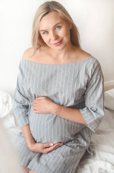 Mujer embarazada feliz sentarse en una cama en casa y mirando a la cámara — Foto de Stock
