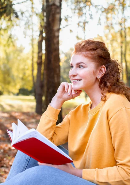 Smiling happy red hair student girl reading a book outside in autumn park