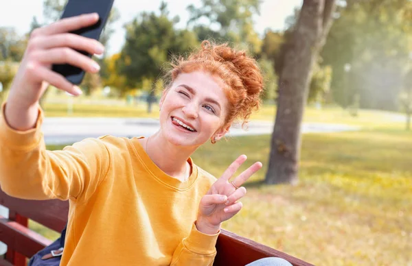 Smiling happy red hair student girl taking picture of herself on the phone outside in autumn park — Stock Photo, Image