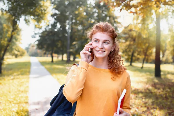 Sorrindo menina estudante cabelo vermelho feliz falando no telefone fora no parque de outono — Fotografia de Stock