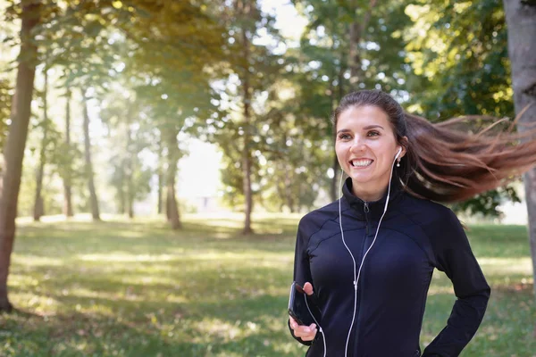 Porträt der jungen lächelnden sportlichen brünetten Frau, die mit Kopfhörern im Park im Freien läuft — Stockfoto