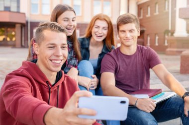 Mutlu gülümseyen bir grup öğrenci üniversitenin yakınındaki açık havada telefonla fotoğraf çekiyor. Eğitim kavramı