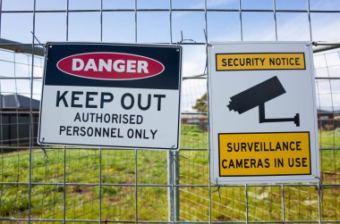 The sign of 'Danger Keep Out Authorised Personnel Only' and ' Security notice Surveillance cameras in use' on a metal construction barrier by a lot of private vacant land. Concept of no trespassing. clipart