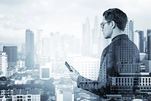 Joven hombre de negocios guapo en traje y gafas usando el teléfono y pensando en cómo abordar el problema, nuevas oportunidades de carrera, MBA. Kuala Lumpur en el fondo. Doble exposición. — Foto de Stock