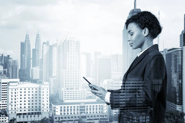 Привлекательная чернокожая афроамериканская деловая женщина в костюме, использующая смартфон и думающая, как решить проблему, новые карьерные возможности, MBA. Куала-Лумпур на заднем плане. Двойное воздействие. — стоковое фото