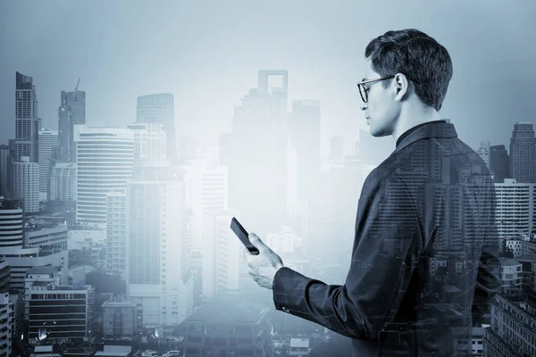 Young handsome businessman in suit and glasses using phone and thinking how to tackle the problem, new career opportunities, MBA. Bangkok on background. Double exposure.