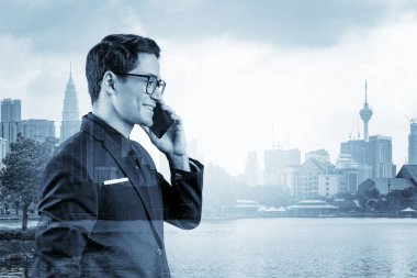 Takım elbiseli, gözlüklü, telefonla konuşan ve Kuala Lumpur şehri manzarasına bakan başarılı yakışıklı girişimci. Sorun çözme danışmanlığı kavramı. KL gökdelenleri. Çift pozlama.