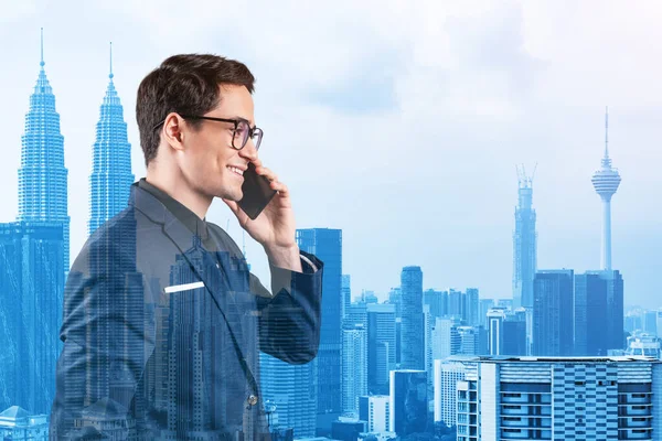 Takım elbiseli, gözlüklü, telefonla konuşan ve Kuala Lumpur şehri manzarasına bakan başarılı yakışıklı girişimci. Sorun çözme danışmanlığı kavramı. KL gökdelenleri. Çift pozlama.