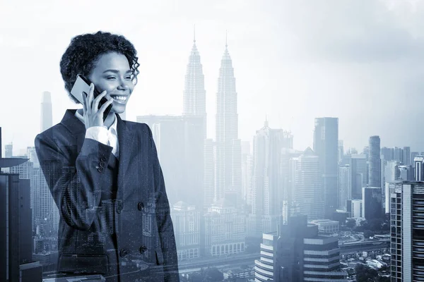 Успішно посміхаючись чорній афроамериканській діловій жінці в костюмі енергійної розмови по телефону, Куала-Лумпур цитайскейп. Концепція консультантів як розв'язувачів проблем. KL skyscrapers Подвійний вплив. — стокове фото