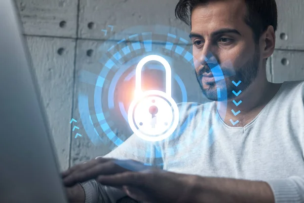 A serious caucasian programmer in casual wears, using laptop to develop a new security system to protect data. Double exposure. IT lock hologram icon.