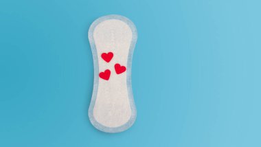 Kan salgılanmasının sembolü olarak kırmızı kalpli, günlük, tek seferlik beyaz bir kadınsı ped. Kadın sağlığı konsepti, adet döngüsü, ICP, nokta. Fotokopi alanı olan mavi düz arkaplan. Hijyenik peçete.