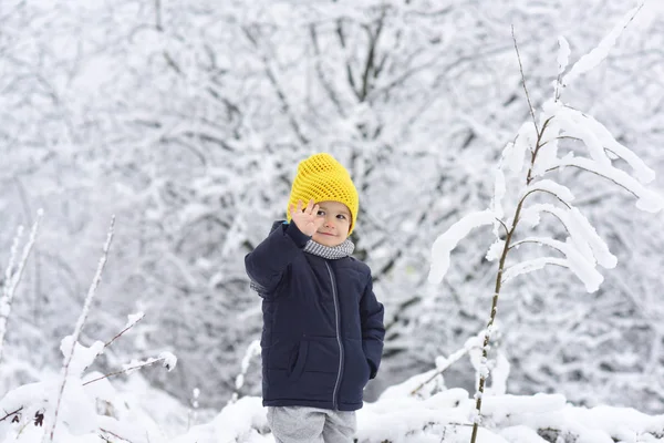 Hello, winter. Handsome boy in black warm jacket and yellow knitted cap. Kid in snow waving hand hello. Friendly boy smiles in the winter forest. Happy childhood. Positive children. Winter walk