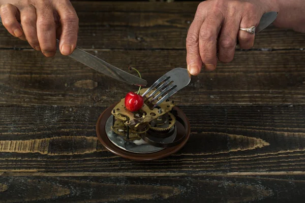 Time concept. Old hands eat clock mechanism for dessert with cherry berry. Wasted time. Mature woman waste time. Lunch time on a wooden background, old age