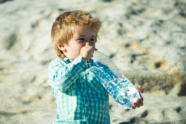 Handsome boy drinking water from big bottle. Thirsty child holding big bottle and tray to drink, hot summer day on beach. Lot of sand background. Thirst and liquid for health in summer. Pure water