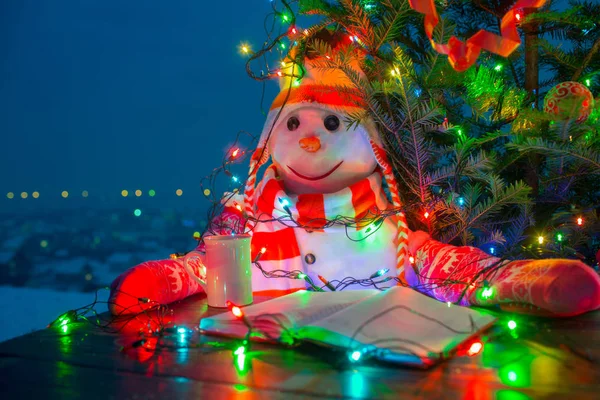 Snowman reading in yard. Education on holidays. Smart holiday time spending. Cozy Christmas with tea, book and Christmas tree. Snowman in red gloves. Work without days off. Exam, test at university