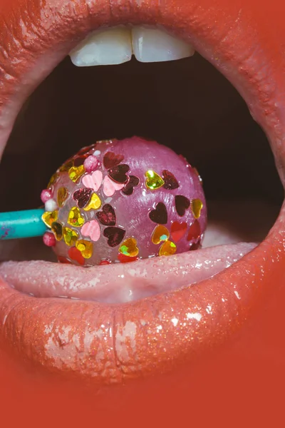 Sexy mouth, tasty tongue. Open female mouth with candy inside. Lollipop covered little hearts. Orange lipstick on lips young beautiful girl. Lip close up with candy. Lip gloss and lipstick