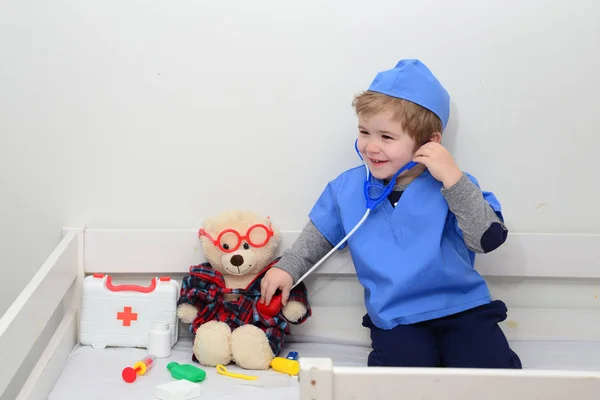 Child playing doctor or nurse with plush toy bear at home. Happy boy listens a stethoscope to toy. Playful boy child playing. Adorable child dressed as doctor playing with toy over white background