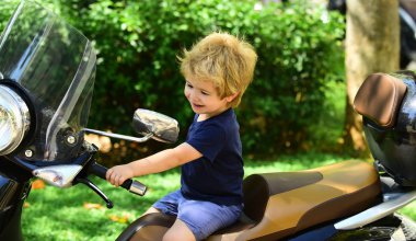 Seyahat. Güzel bir çocuk mavi T-shirt motosiklet oturur. Küçük çocuk. Temiz hava sürme motosiklet. Zemin üzerine yeşil çalılar. Genç Sarışın motosikletçi yürüyüş.