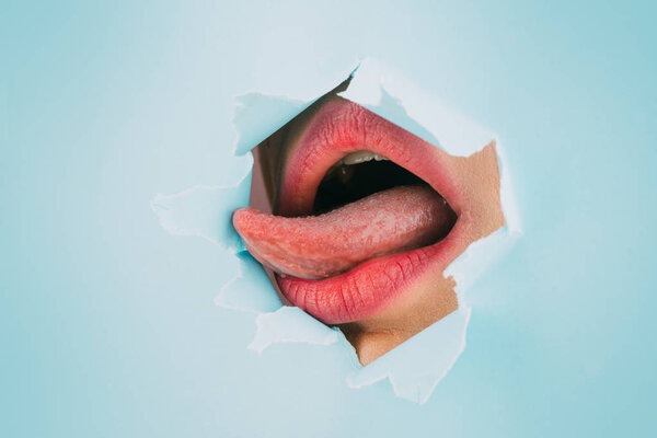 Womans open mouth with sexy pink lips and tongue. Fashion photography. Natural full lips with bright lip makeup. Pink lipstick and lip gloss in a paper hole, blue background.