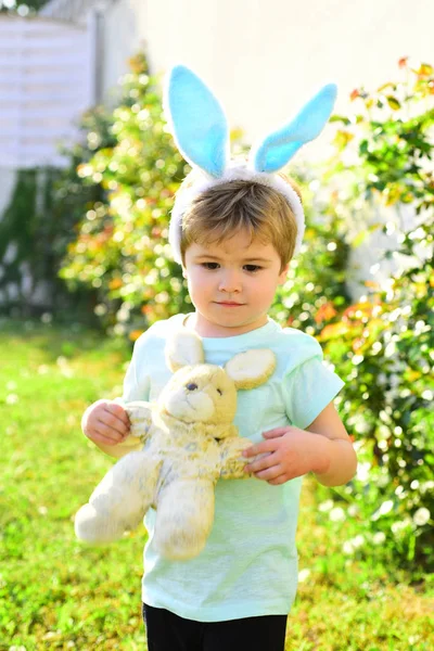 Das kleine Kind verbringt Zeit mit seinem Teddyfreund draußen. Sommerurlaub mit der Familie auf dem Land. Porträt eines konzentrierten Jungen im Garten mit Teddyspielzeug und großen Hasenohren. Ostern — Stockfoto
