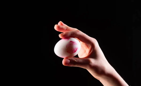 Egg kiss. Lip print from red lipstick on chicken egg. Creative Easter. Female hand holding egg. Symbol of life on a black background.