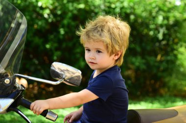 Motosiklet arka görüş aynası. Yalnız çocuk sıcak yaz gününde gümüş büyük motosiklet üzerinde oturur. Dışarıda küçük bir motosikletçi var. Sevimli çocuk mutlu duygular. Arka planda doğa. Yeşil çalılar ve büyük motosiklet.
