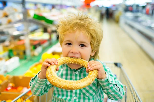 Uma criança esfomeada no supermercado. Menino loiro bonito senta-se no carrinho de compras e come pão redondo com sementes de tempero. Um miúdo independente sozinho na loja. Pão fresco. O apetite do bebé. Comida para crianças pequenas — Fotografia de Stock