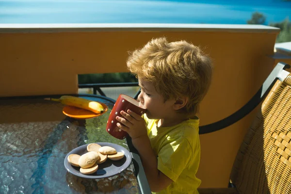 リラックス。バルコニーに座り、新鮮な空気を楽しみ、クッキーとミルクを飲む少年。一人で海の近くの夏休みに子供のカラフルな明るい写真。子供との家族の休日。健康的で速いスナック. — ストック写真