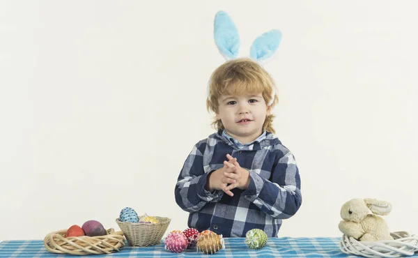 Kind mit Hasenohren auf dem Kopf spielt mit verzierten Ostereiern. Spiele für Kinder in jungen Jahren. Tisch mit blau-weißer Tischdecke und flauschigem Kaninchen im Vordergrund — Stockfoto